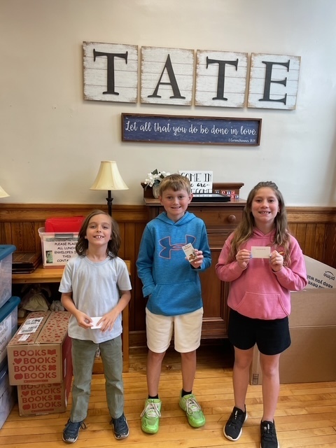 Three kids standing in front of a sign that reads TATE holding chick fil a gift cards