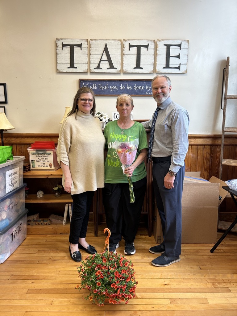 A man and two women standing in front of a sign that says TATE. There is a basket of red flowers in front of them.