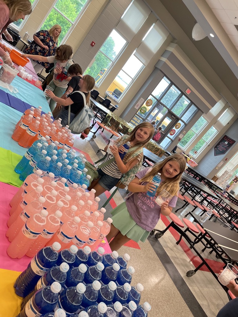 Kids lined up besied a table with colorful tablecloths. Clear plastic cups filled with ice , and bottled waters are sitting on the tables., the kids are holding a clear cup and water bottle.