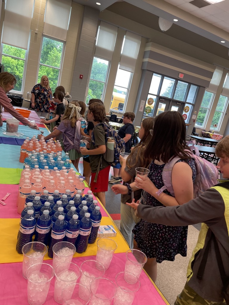 Kids lined up besied a table with colorful tablecloths. Clear plastic cups filled with ice , and bottled waters are sitting on the tables., the kids are holding a clear cup and water bottle.
