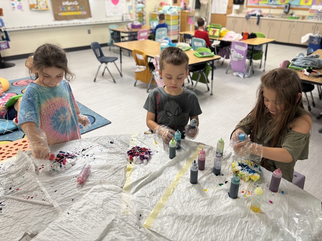 Three children wearing plastic gloves, holding bottles of dye, trash bags are on the table along with more bottles of dye.