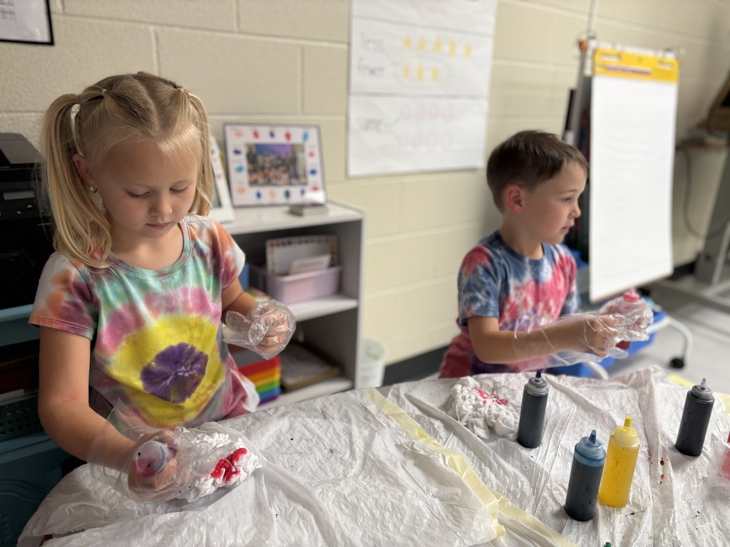 Two small children wearing tie dye shirts. They are wearing plastic gloves, trash bags are on the table along with bottles of dye.