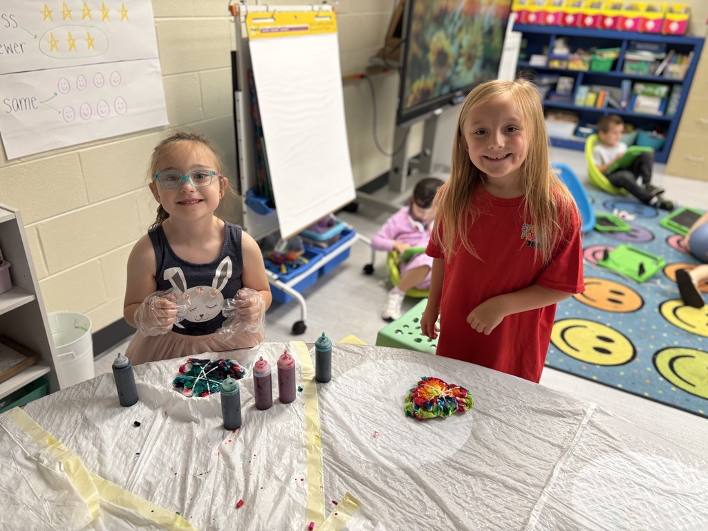 Two children, one is wearing plastic gloves trash bags are on the table along with more bottles of dye. Another child is standing beside her with her finished tie dye shirt wrapped up.
