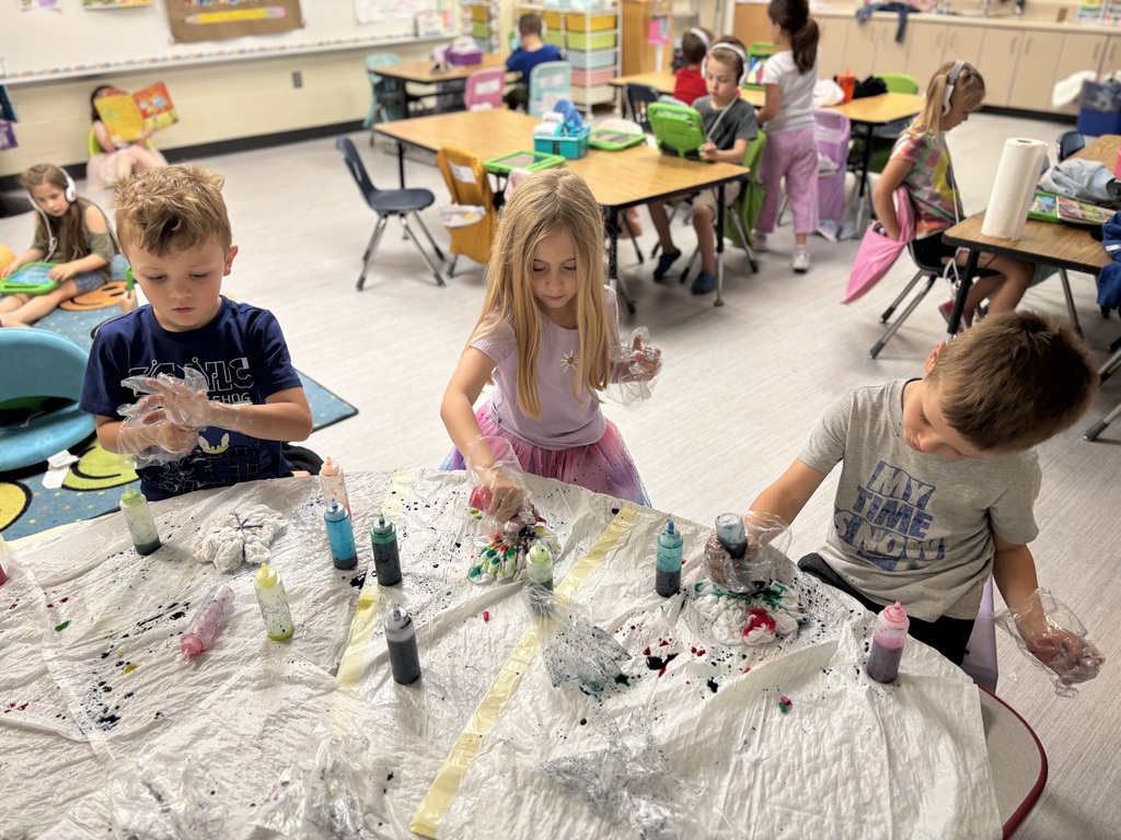 Three children wearing plastic gloves, holding bottles of dye, trash bags are on the table along with more bottles of dye.