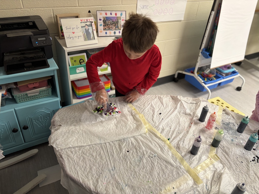One child wearing plastic gloves, holding a bottle of dye, trash bags are on the table along with more bottles of dye.