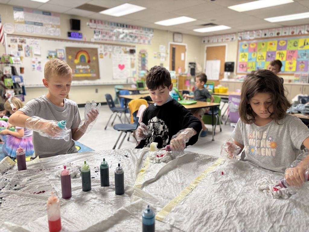 Three children wearing plastic gloves, holding bottles of dye, trash bags are on the table along with more bottles of dye.