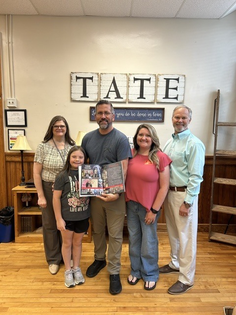four adults and one child standing in front of a sign that says tate. A man iin the middle of the picture is holding a yearbook.