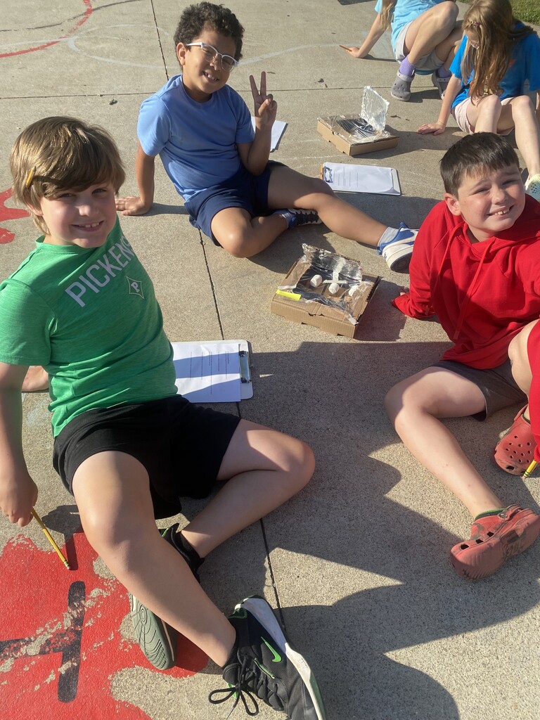 Three female students sitting on the concrete with clipboards and a box on the ground.