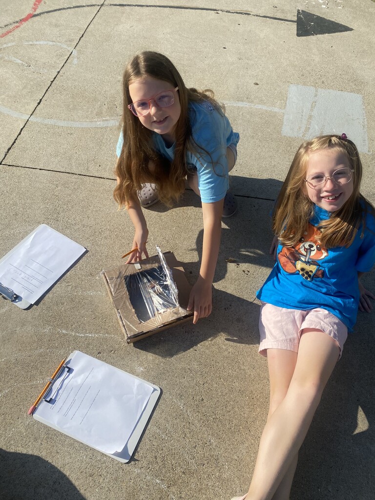 Two female students sitting on the concrete with clipboards and a box on the ground.