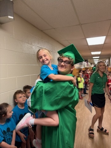 Male High school student wearing a green cap and gown holding a female child. Children are in the background lined up in the hallway
