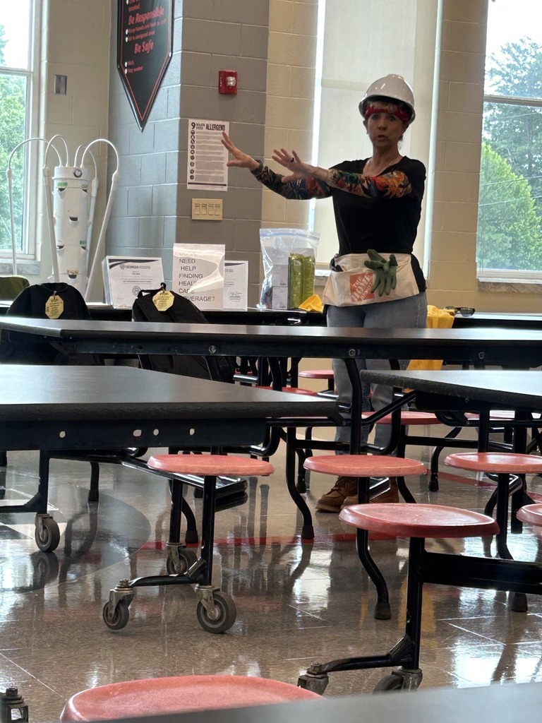  Woman with a white hardhat, black shirt, jeans, and arm tattoo sleeves standing in a school cafeteria with her arms raised