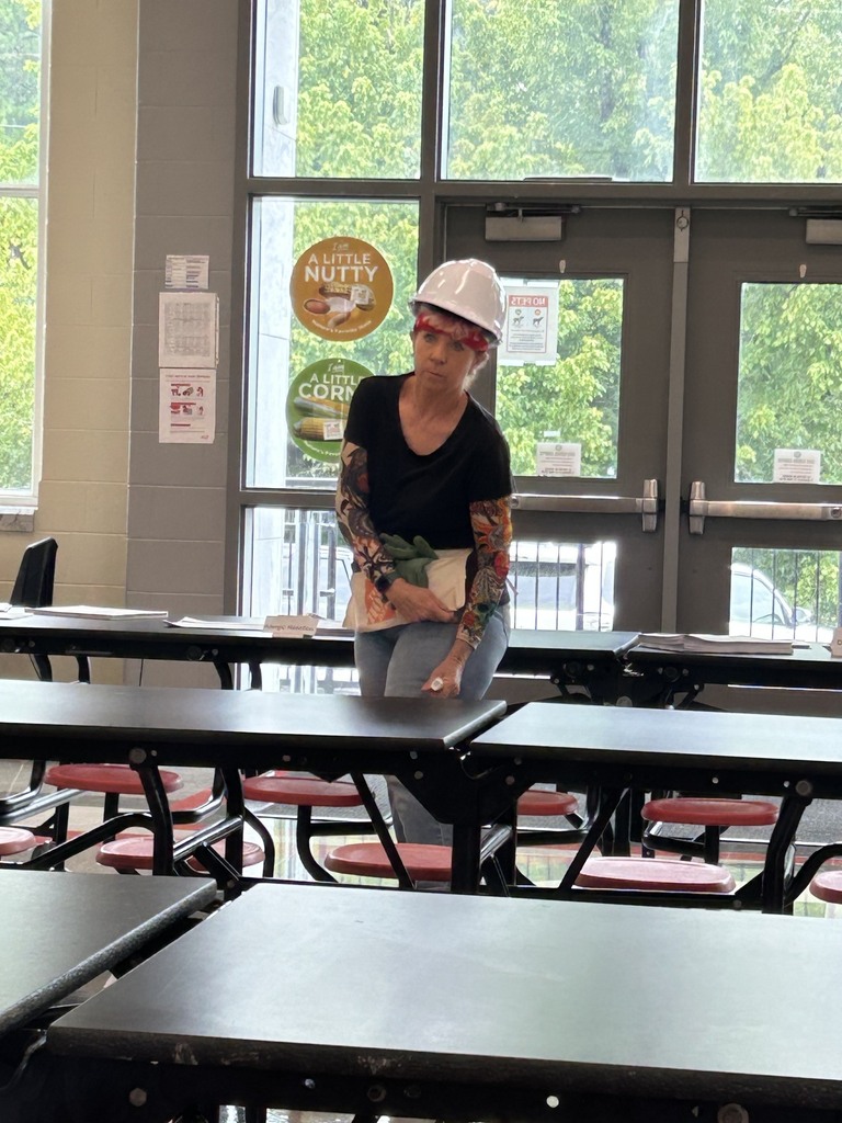  Woman with a white hardhat, black shirt, jeans, and arm tattoo sleeves standing in a school cafeteria using an epipen