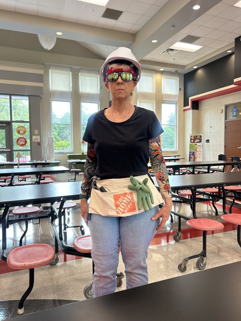 Woman with a white hardhat, black shirt, jeans, and arm tattoo sleeves standing in a school cafeteria