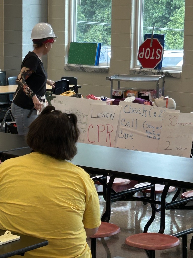  Woman with a white hardhat, black shirt, jeans, and arm tattoo sleeves standing in a school cafeteria, woman in yellow shirt sitting at the cafeteria table