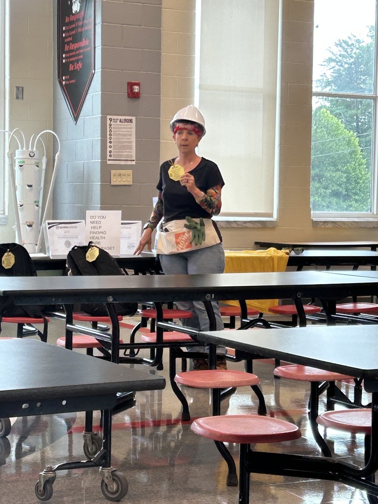  Woman with a white hardhat, black shirt, jeans, and arm tattoo sleeves standing in a school cafeteria holding a yellow sign