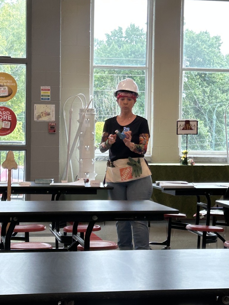  Woman with a white hardhat, black shirt, jeans, and arm tattoo sleeves standing in a school cafeteria using an inhaler
