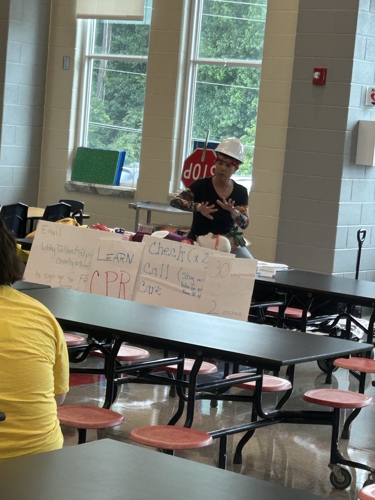  Woman with a white hardhat, black shirt, jeans, and arm tattoo sleeves sitting and doing compressions on a cpr dummy in a school cafeteria
