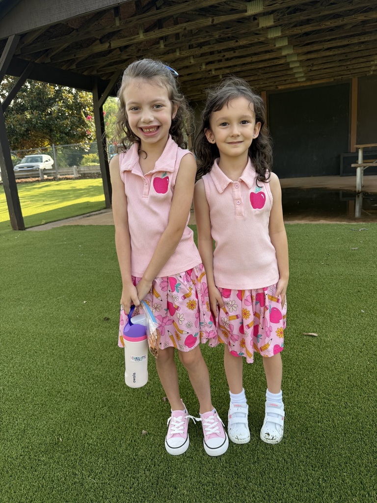 Two female students wearing matching clothes of light pink with an apple on the front pocket. They are standing on the playground.