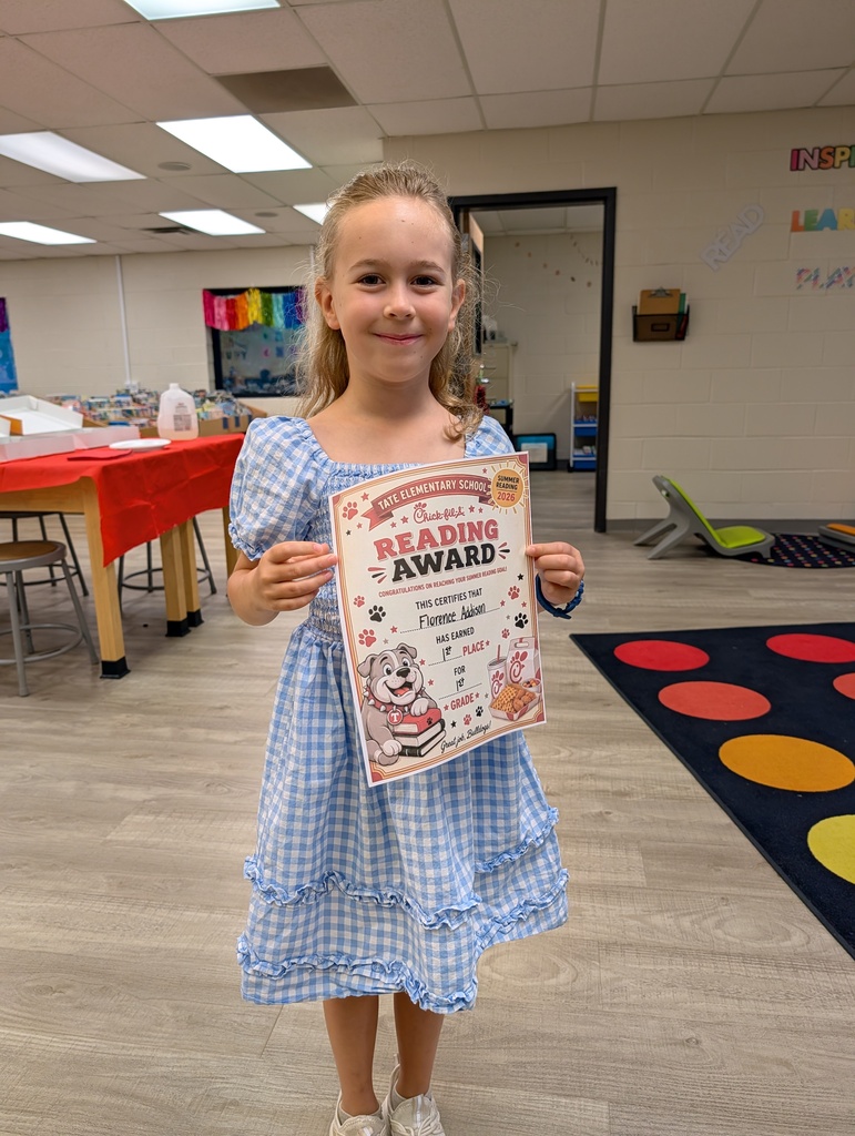 A student stands in a school library holding a “Reading Award” certificate toward the camera, smiling, with other tables and a colorful wall sign and carpet in the background.