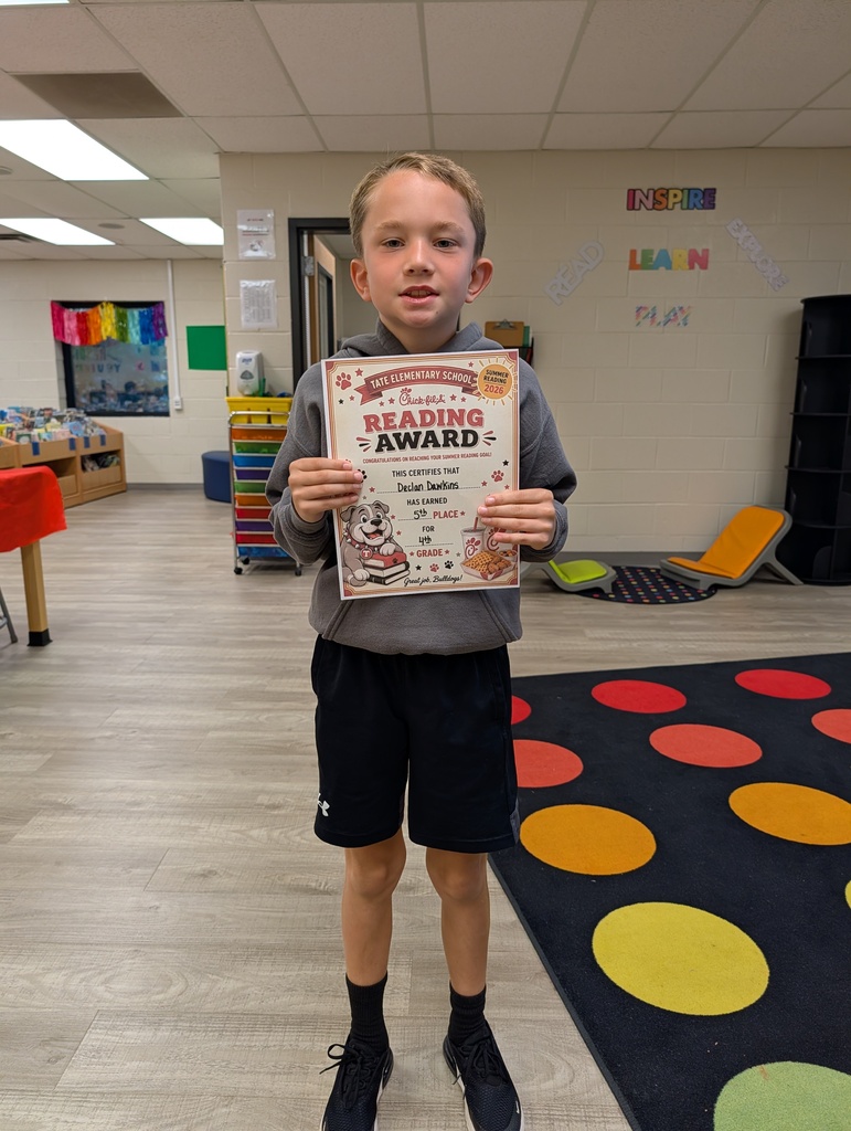 A student stands in a school library holding a “Reading Award” certificate toward the camera, smiling, with other tables and a colorful wall sign and carpet in the background.