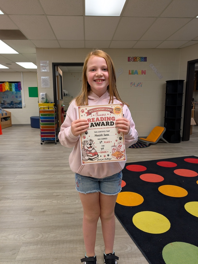 A student stands in a school library holding a “Reading Award” certificate toward the camera, smiling, with other tables and a colorful wall sign and carpet in the background.