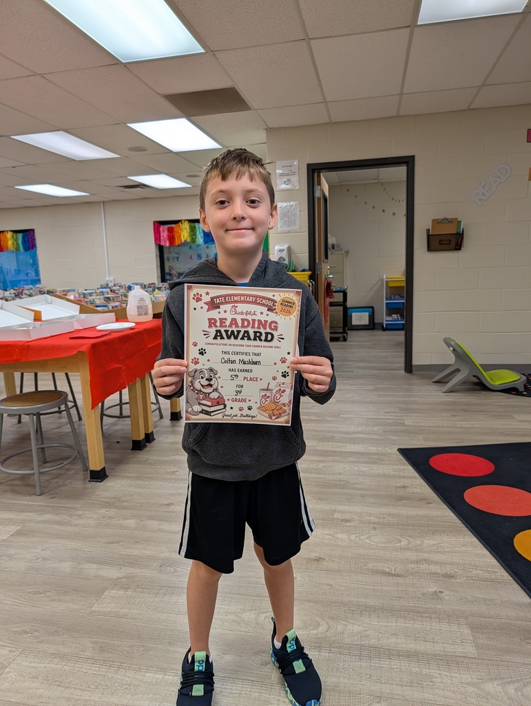 A student stands in a school library holding a “Reading Award” certificate toward the camera, smiling, with other tables and a colorful wall sign and carpet in the background.