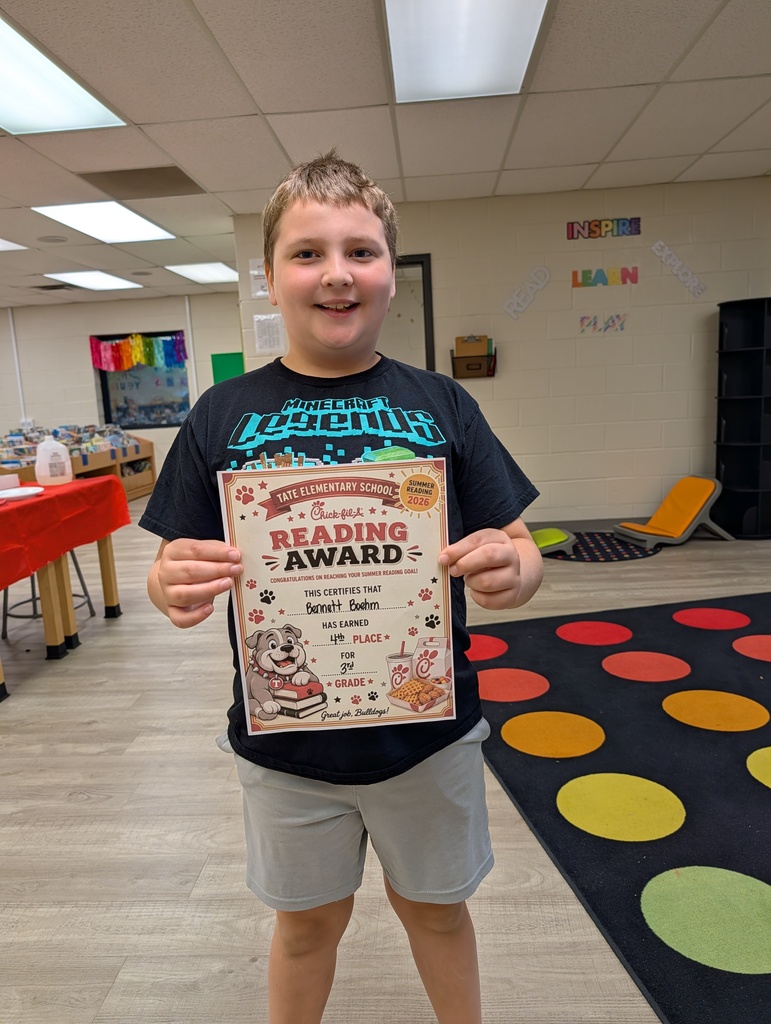 A student stands in a school library holding a “Reading Award” certificate toward the camera, smiling, with other tables and a colorful wall sign and carpet in the background.