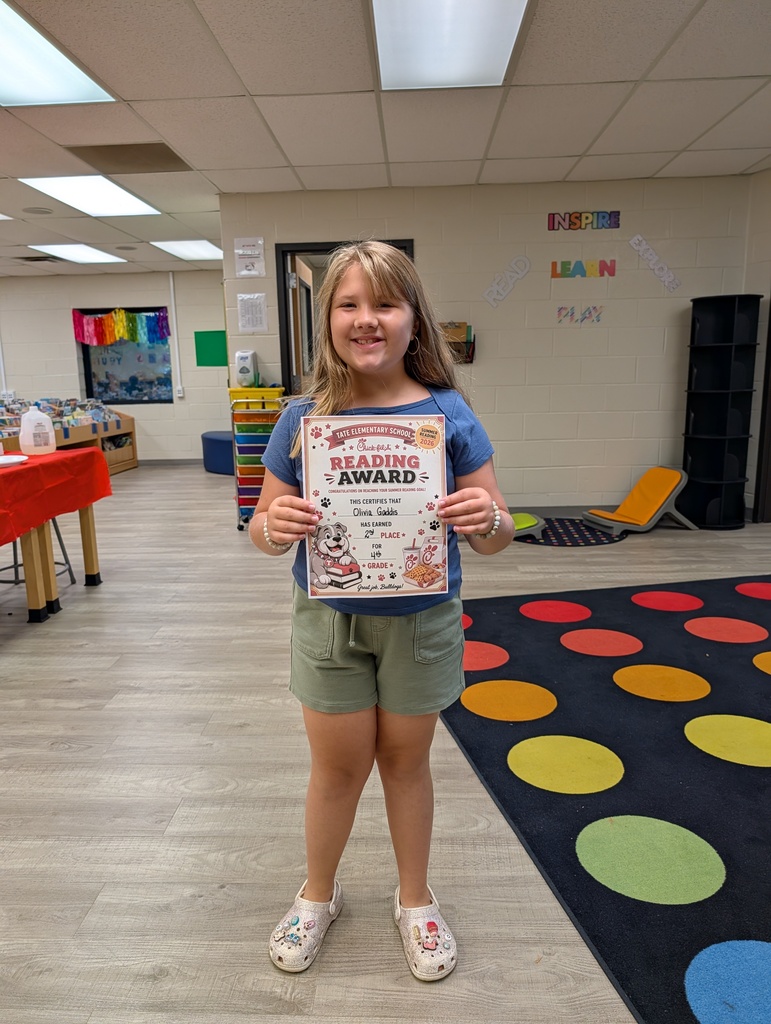 A student stands in a school library holding a “Reading Award” certificate toward the camera, smiling, with other tables and a colorful wall sign and carpet in the background.