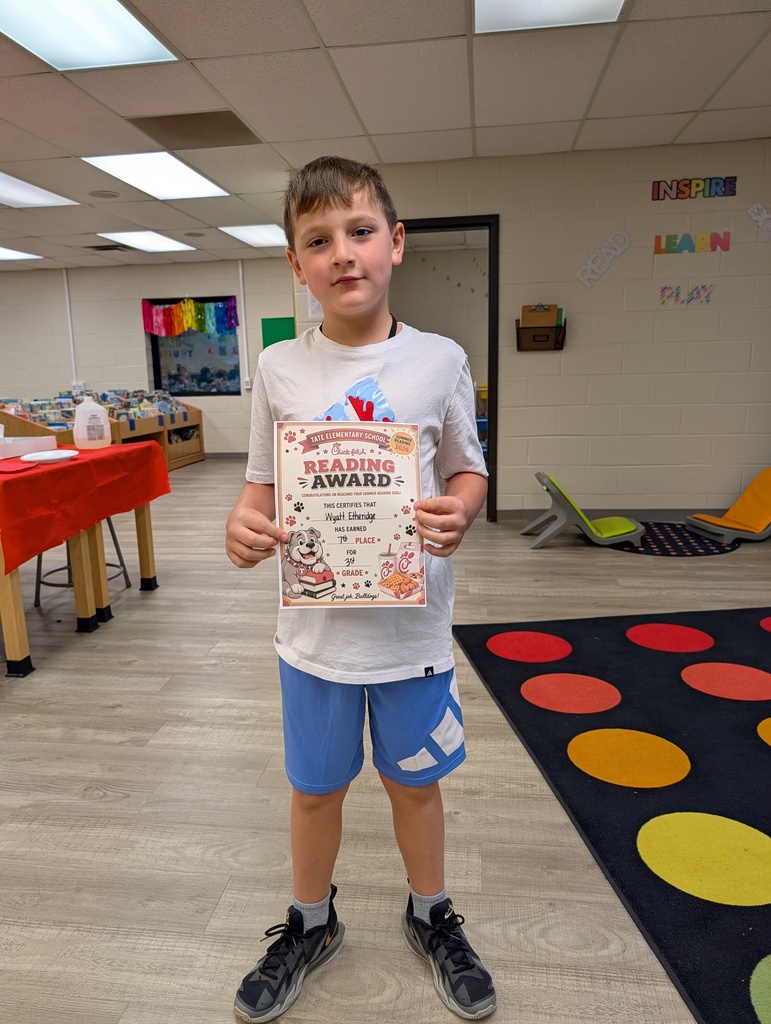 A student stands in a school library holding a “Reading Award” certificate toward the camera, smiling, with other tables and a colorful wall sign and carpet in the background.