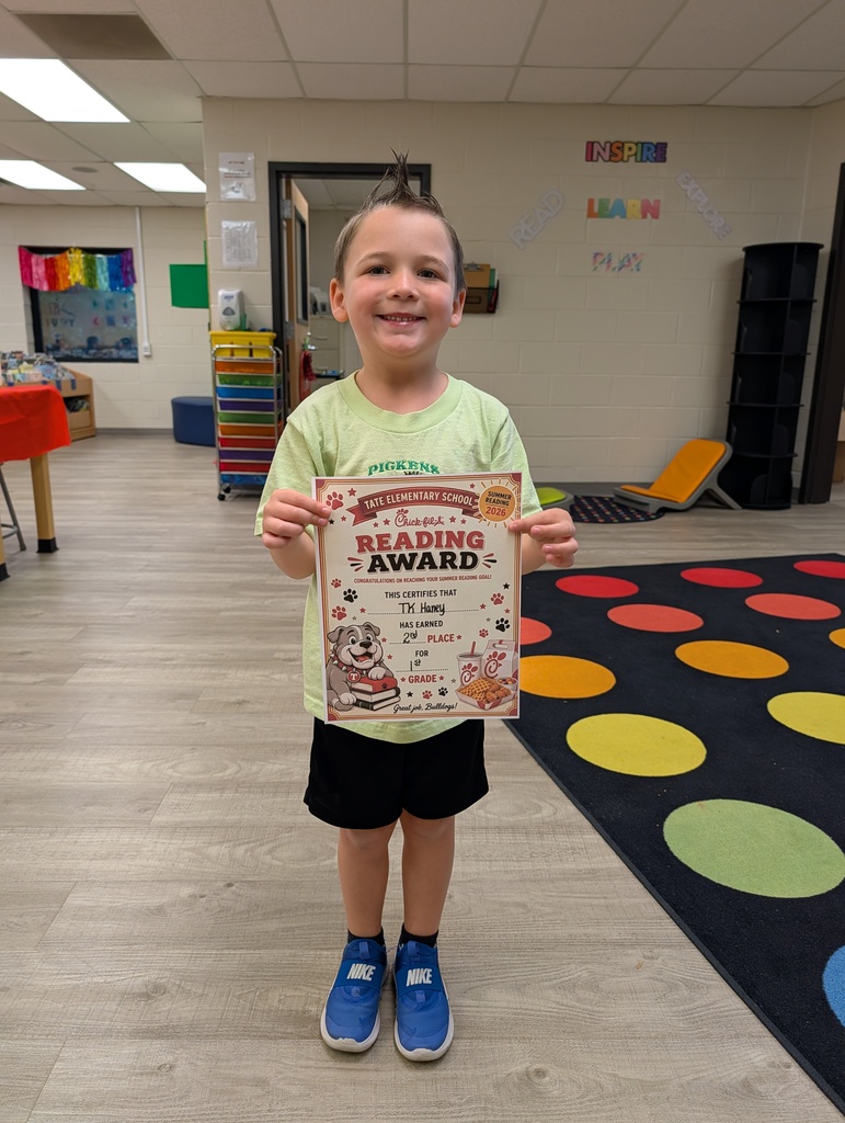A student stands in a school library holding a “Reading Award” certificate toward the camera, smiling, with other tables and a colorful wall sign and carpet in the background.