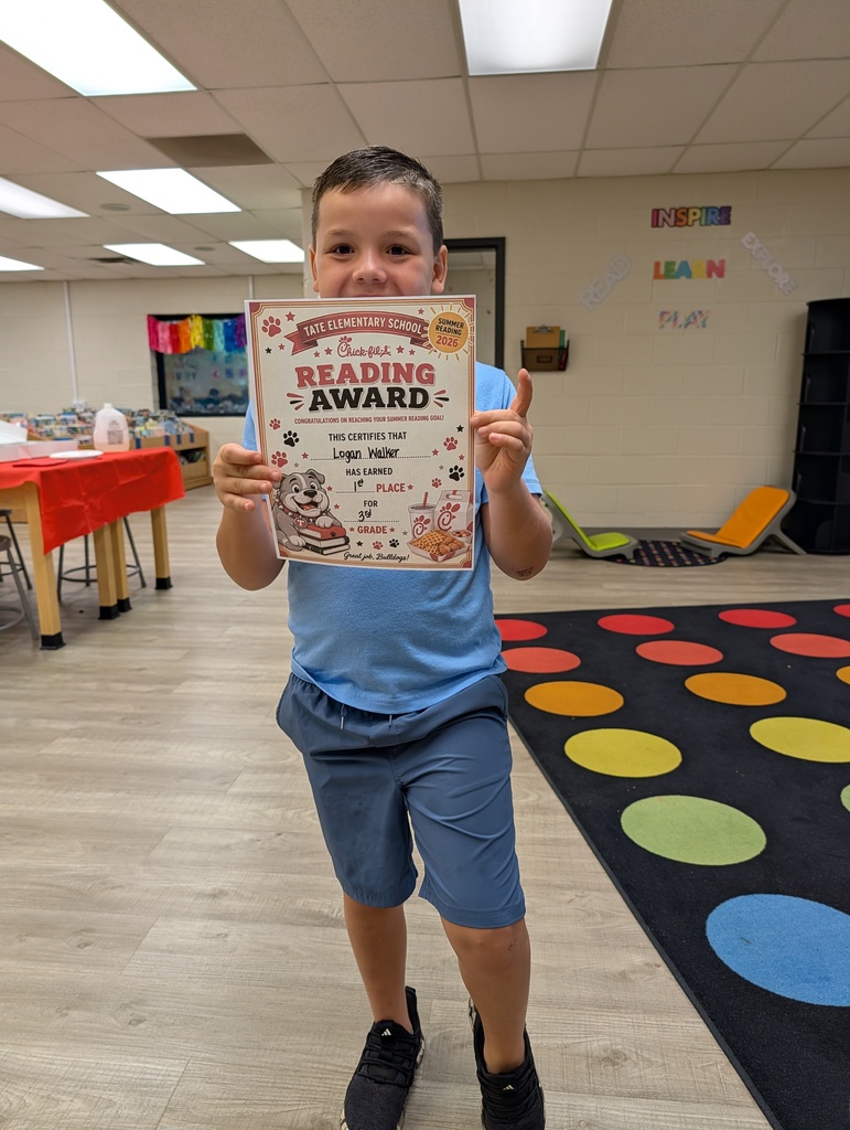 A student stands in a school library holding a “Reading Award” certificate toward the camera, smiling, with other tables and a colorful wall sign and carpet in the background.