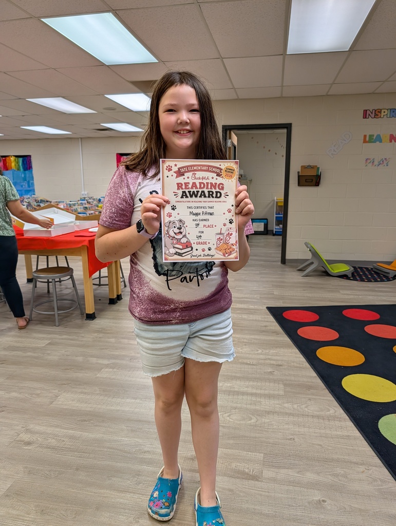 A student stands in a school library holding a “Reading Award” certificate toward the camera, smiling, with other tables and a colorful wall sign and carpet in the background.