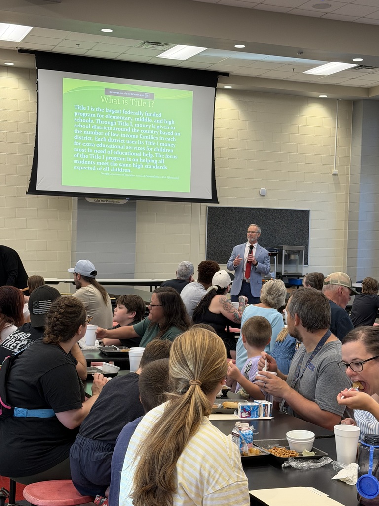A speaker stands in front of a projected slide titled “What is Title 1?” while an audience sits at tables with food and drinks, listening and looking toward the screen.
