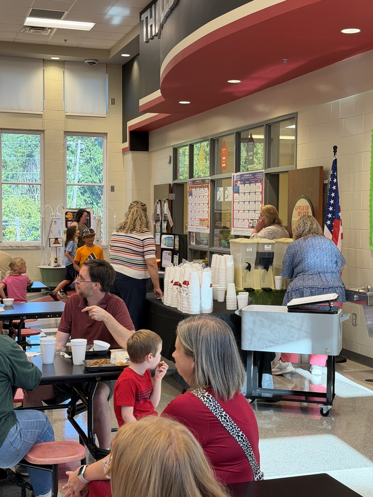 People seated at tables inside a school cafeteria, watching a presenter near a service counter with cups, paper items, and a U.S. flag, with windows and bulletin boards along the wall.