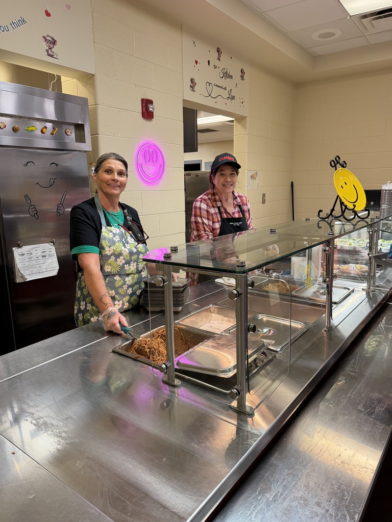 Two people stand behind a stainless-steel food service counter in a brightly lit cafeteria, smiling while preparing items behind a glass display case with colorful menu drawings on the wall.