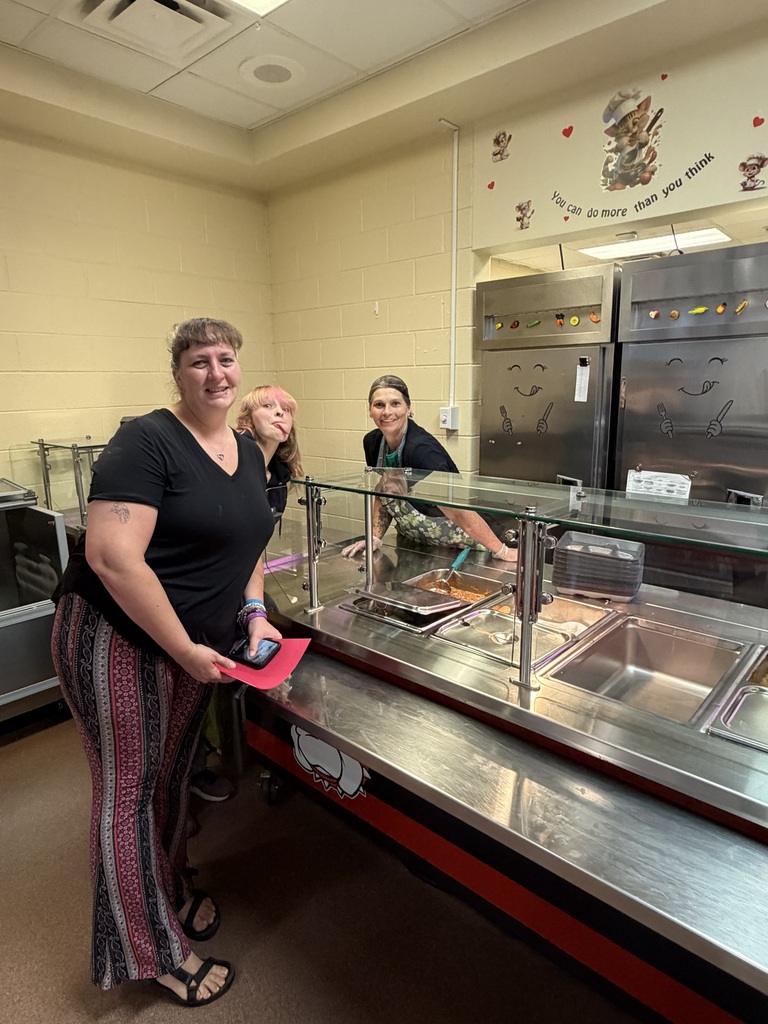 In a school cafeteria kitchen, a woman stands by a stainless-steel food service counter while others work behind the glass; warm lighting, tiled walls, and red heart wall decals are visible.