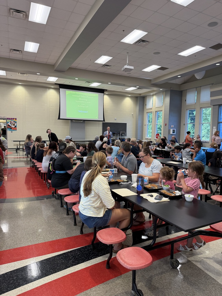 A presenter stands at the front of a school cafeteria speaking to an audience seated at tables while a projector displays a green-tinted slide titled “What is Title I?” on a screen.