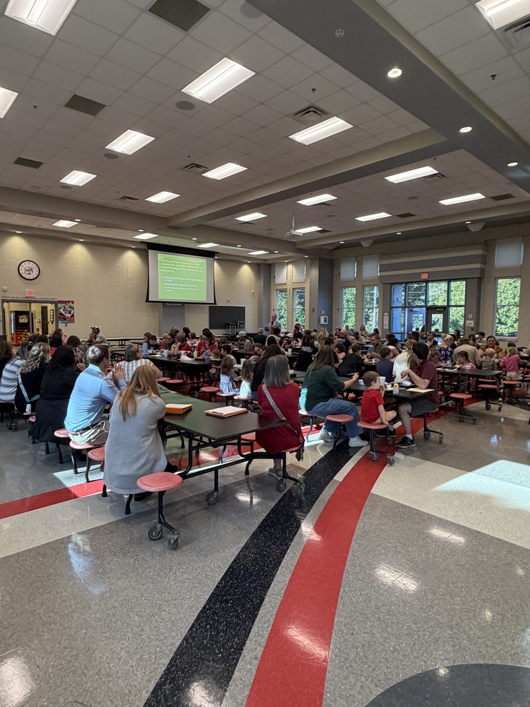 A school cafeteria filled with students and adults seated at long tables, facing a front screen with a presentation, while a teacher stands near the board under bright ceiling lights.