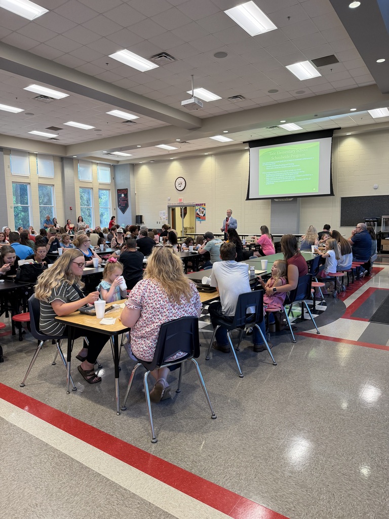A school cafeteria filled with adults and students seated at tables, listening as a presenter speaks at the front beside a large projector screen with a green slide.