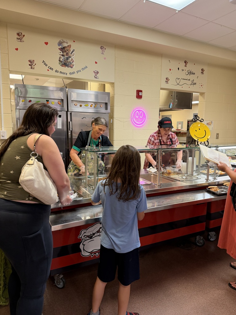 People stand at a school-style ice cream counter with glass cases and red-and-black trim, while staff serve behind the counter; a child waits in front and colorful wall decals add playful decorations.