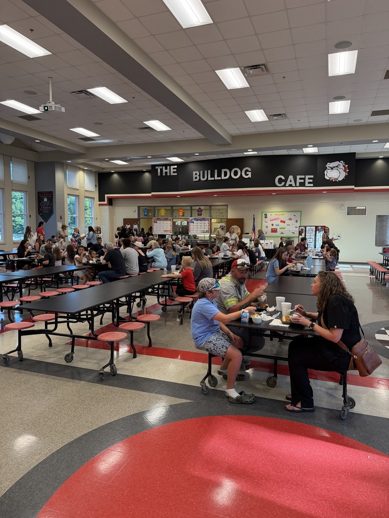 A busy school cafeteria with many students and adults seated at tables and eating, red and black chairs, and a “THE BULLDOG CAFE” sign on the back wall under bright ceiling lights.