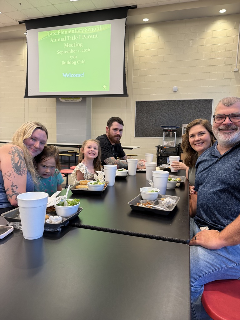 Family gathered around a cafeteria table with white cups and bowls during a school welcome event; a projector screen in the background reads “Annual Title I Parent Meeting” and “Welcome!”
