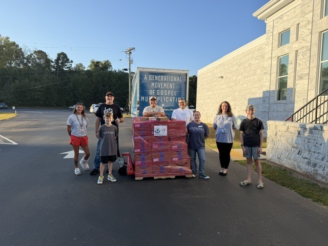 A group of people standing in a parking lot beside a large box trailer packed with red-and-white items, with a building on the right and a banner reading “A Generational Movement of Gospel…” in the background.
