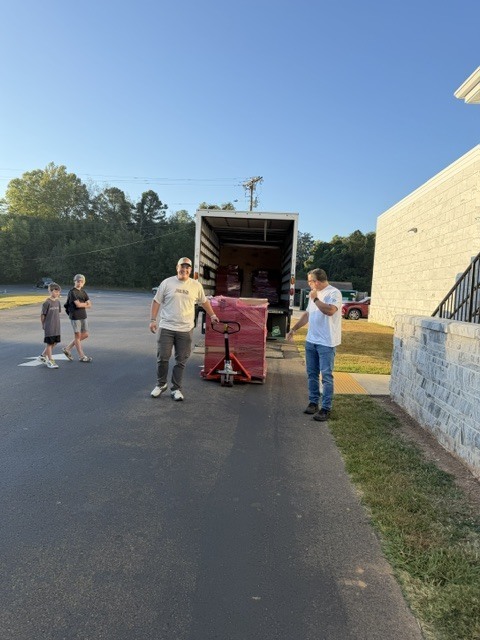  group of three people stand on a paved driveway beside a red-and-black grill or smoker unit, with a large open trailer behind them and trees and a clear blue sky in the background.