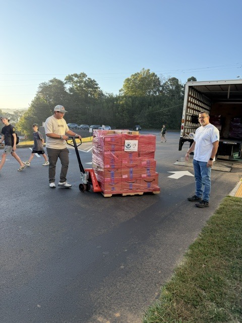 A group of people walk along a paved driveway while a large pallet of red-wrapped boxes sits on a cart in front of them near an open box truck.