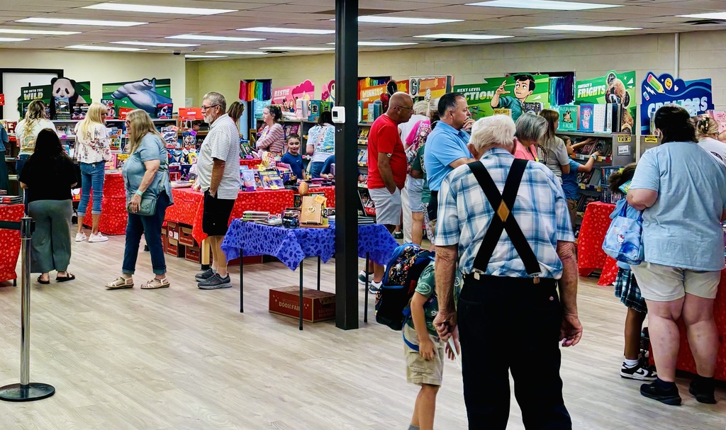 People browse shelves of books and toys in a brightly lit school media center, with colorful displays and gift items on tables and racks, seen from behind near a central pillar.