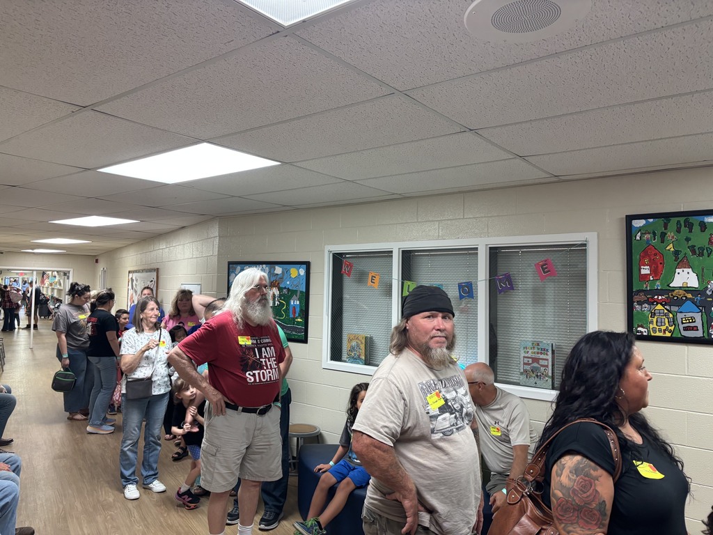 People line a school hallway near classroom doors decorated with colorful posters; a long-bearded man in a red shirt and a man in a gray hat stand among others while students watch and wait.