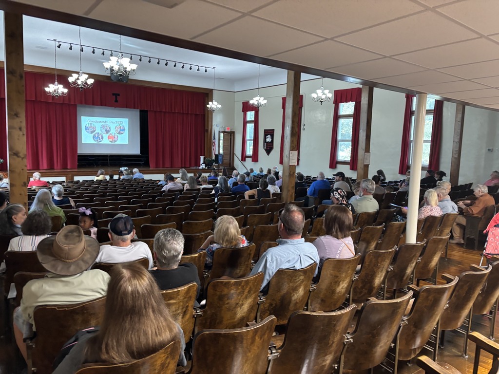 An audience sits in wooden auditorium seats facing a stage with red curtains and a large screen showing a presentation.