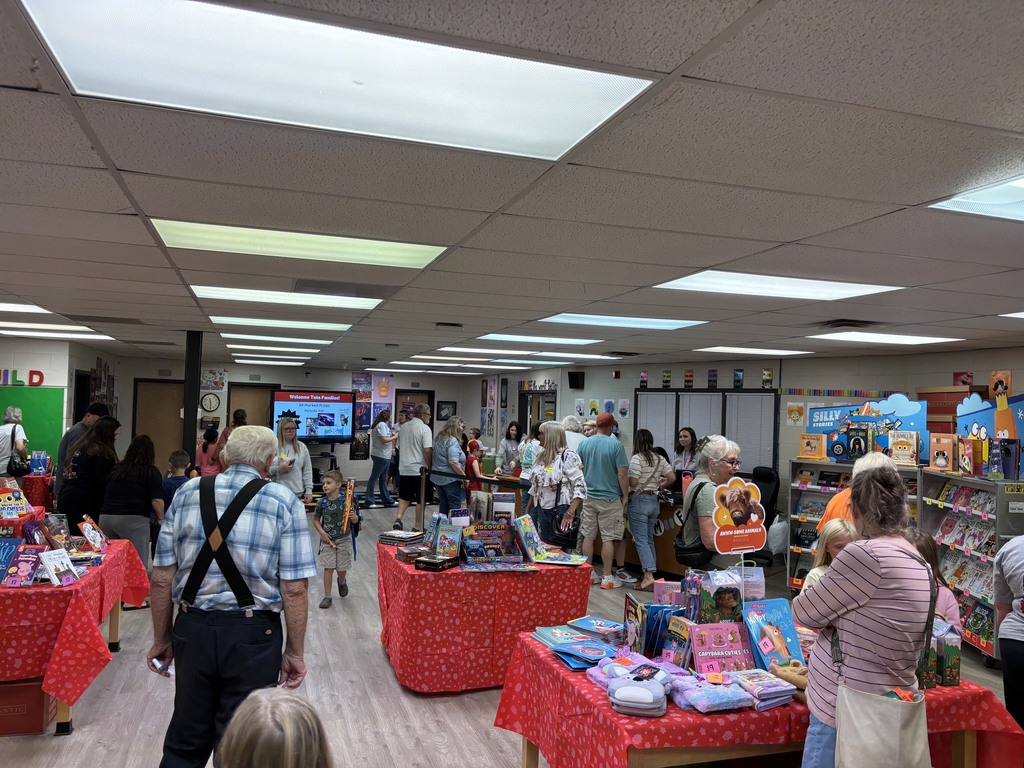 Busy indoor bookfair in a school Media Center, with people browsing tables covered in colorful toys and books also on displays under bright ceiling lights.