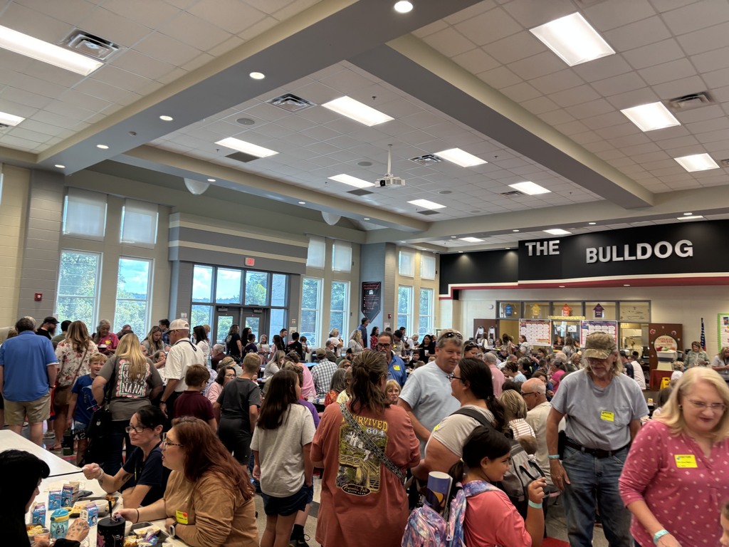 Crowd gathered inside a school cafeteria with a “THE BULLDOG CAFE” sign above serving counters, people in line near red flooring and tables, and posters on the walls.